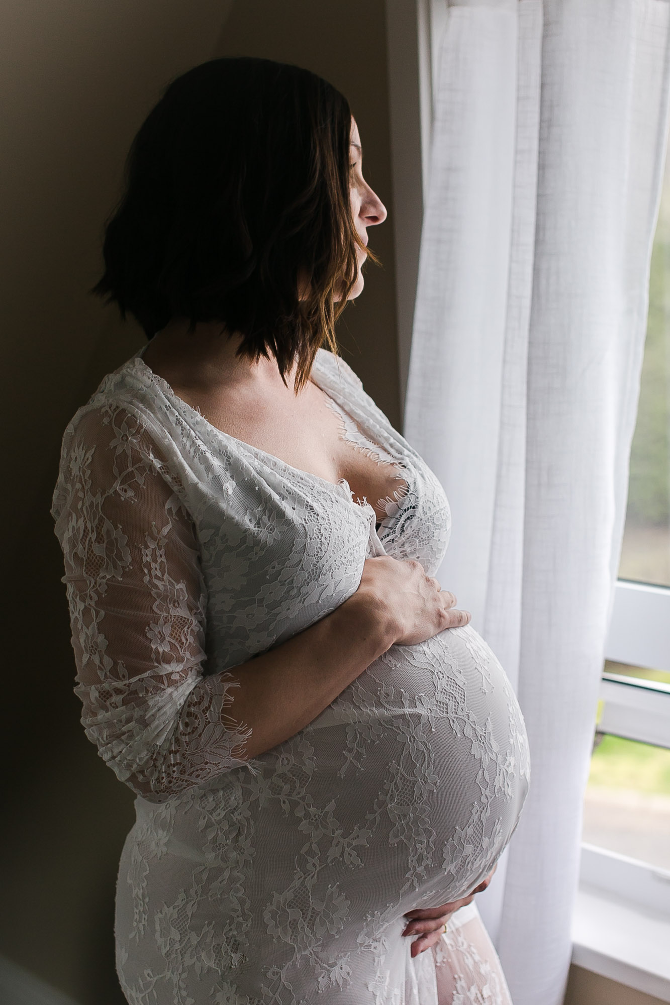 New Westminster maternity photo session Pregnant woman stands by a window in a lace dress during an in-home lifestyle maternity session.